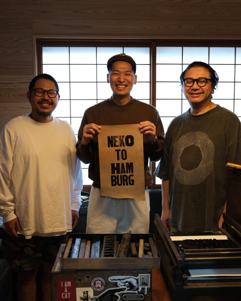 NINE LETTERPRESSの活版印刷ワークショップで、木活字を使って制作したNEKOTOHAMBURGの印刷物を囲む参加者3人